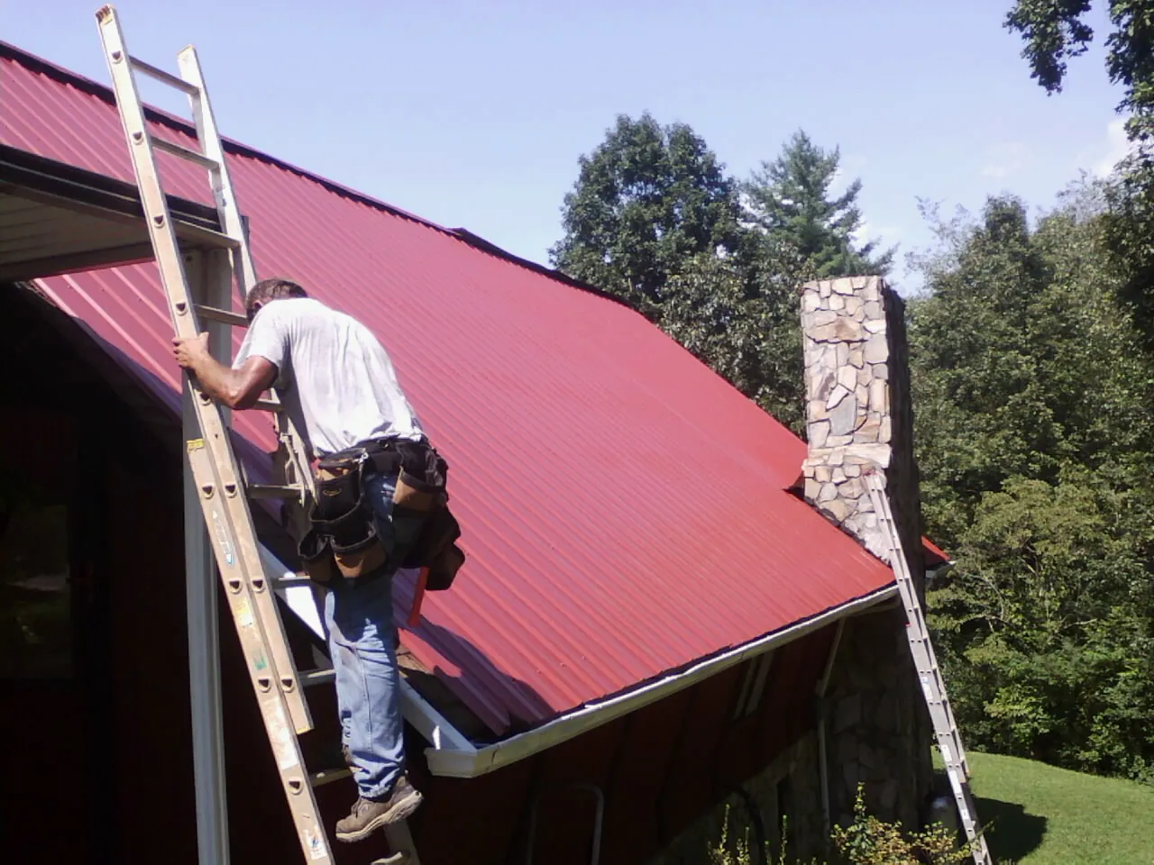 Roofer on a ladder beside a red metal roof and stone chimney — Soffit & Fascia Repair by Roofing Brothers in West Milton
